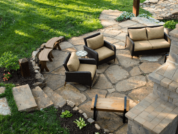 Backyard patio with natural flagstone flooring, featuring cushioned wicker lounge chairs, a wooden coffee table, and curved wooden benches around a fire pit. The space is bordered by lush green grass, small garden beds, and stone steps leading into the area, creating a cozy and inviting outdoor living space.